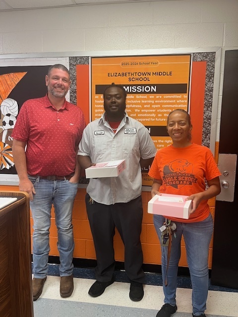 Three Elizabethtown Middle School staff members stand smiling in front of an orange and black mission statement wall, holding gift boxes in recognition of Custodian Appreciation Day.