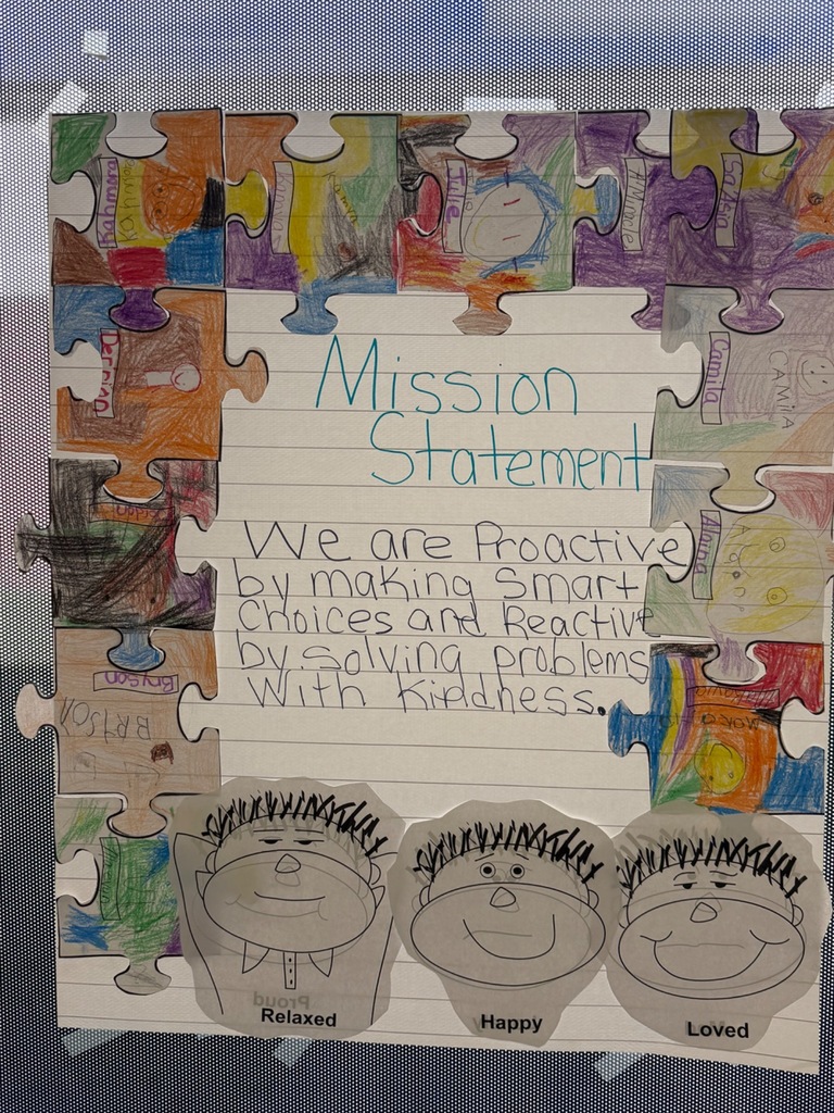A colorful classroom mission statement is displayed on lined paper surrounded by puzzle pieces decorated and colored by students. The center reads, “Mission Statement: We are proactive by making smart choices and reactive by solving problems with kindness.” Below the writing are three cartoon faces labeled “Relaxed,” “Happy,” and “Loved.” The border puzzle pieces include students’ names and drawings in bright colors, showing creativity and teamwork.