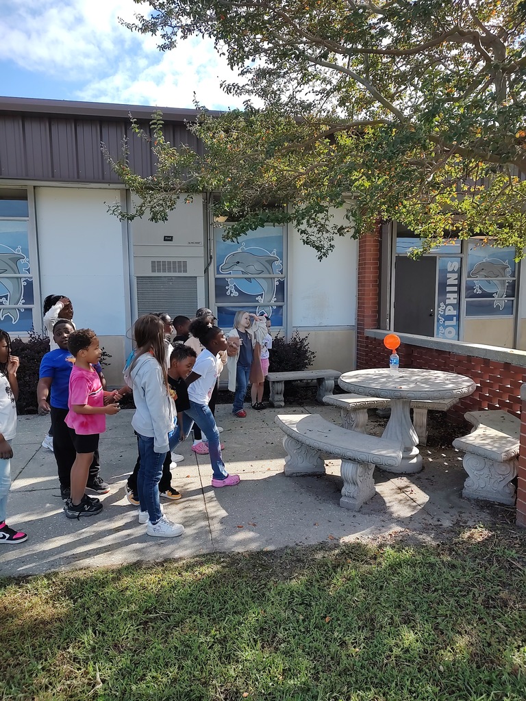 Students stand outside near a concrete picnic table observing an orange balloon attached to a bottle in the sunlight. They appear engaged as they watch a science experiment take place, with school buildings and dolphin-themed window art in the background.