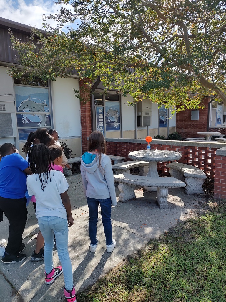 Students stand outside near a concrete picnic table observing an orange balloon attached to a bottle in the sunlight. They appear engaged as they watch a science experiment take place, with school buildings and dolphin-themed window art in the background.