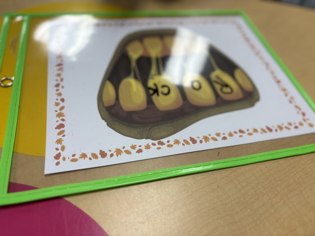 A child practices reading skills using a laminated sheet decorated with fall leaves that shows a monster’s mouth and teeth. The student writes letters on the monster’s teeth and uses a blue toothbrush to “brush” across them while blending sounds to form words.