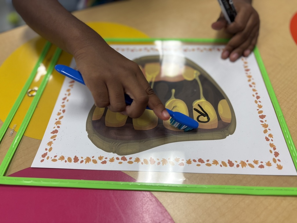 A child practices reading skills using a laminated sheet decorated with fall leaves that shows a monster’s mouth and teeth. The student writes letters on the monster’s teeth and uses a blue toothbrush to “brush” across them while blending sounds to form words.