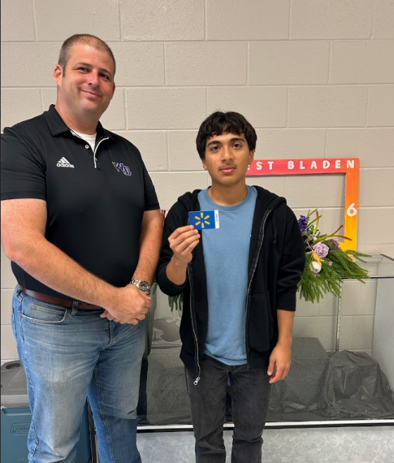 A West Bladen staff member stands beside a student holding a Walmart gift card. Both are smiling slightly while posing against a gray brick wall with a “West Bladen” sign and flower arrangement in the background.