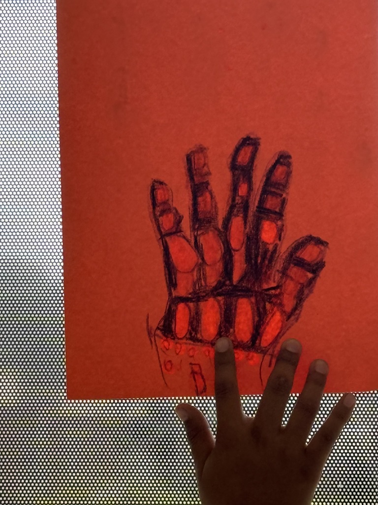 A child's hand reaches toward a red sheet of paper displaying a drawn hand, illuminated by light through a perforated surface.