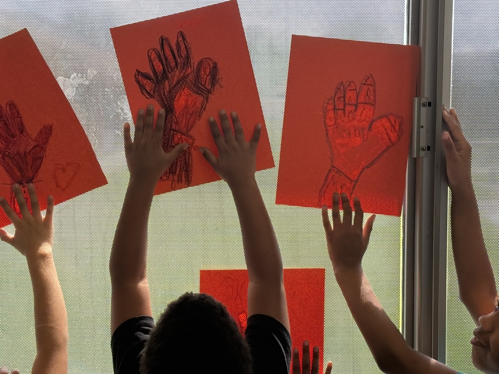 Children's hands reaching up to red posters of hand drawings, displayed against a light-filtering window.