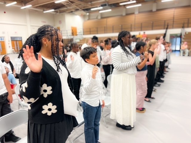 Students dressed in black and white raise their right hands during a ceremony in a school gymnasium.
