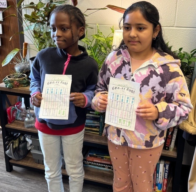 Two elementary students proudly hold up papers labeled “Great Job!” while standing in front of a bookshelf and plants.