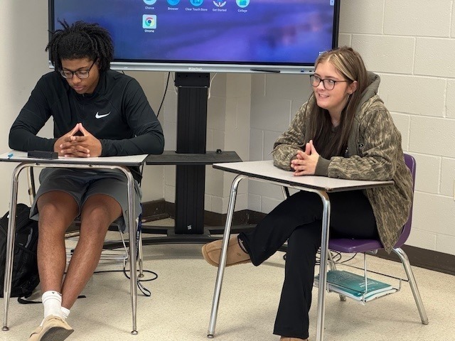 Two high school students sit at desks during a class discussion, one smiling and the other looking down thoughtfully.