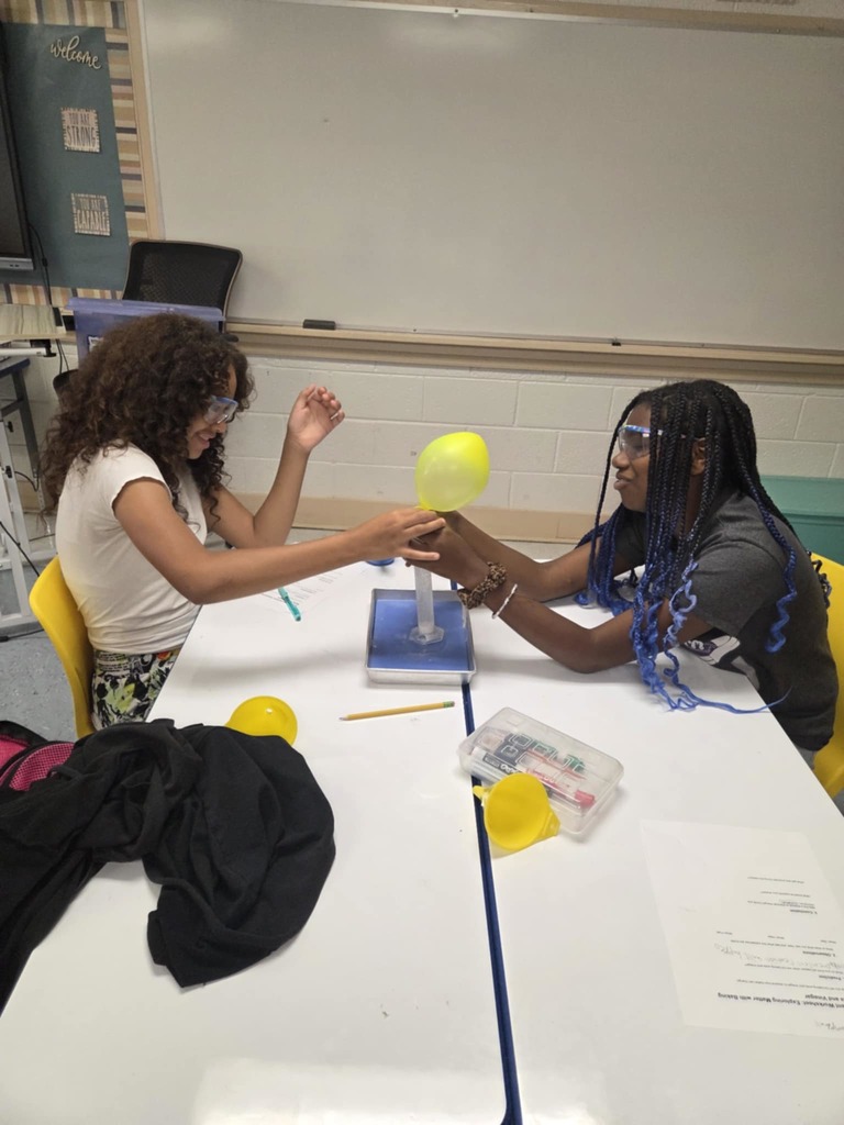 Two students wearing safety goggles work together on a science experiment involving a yellow balloon.