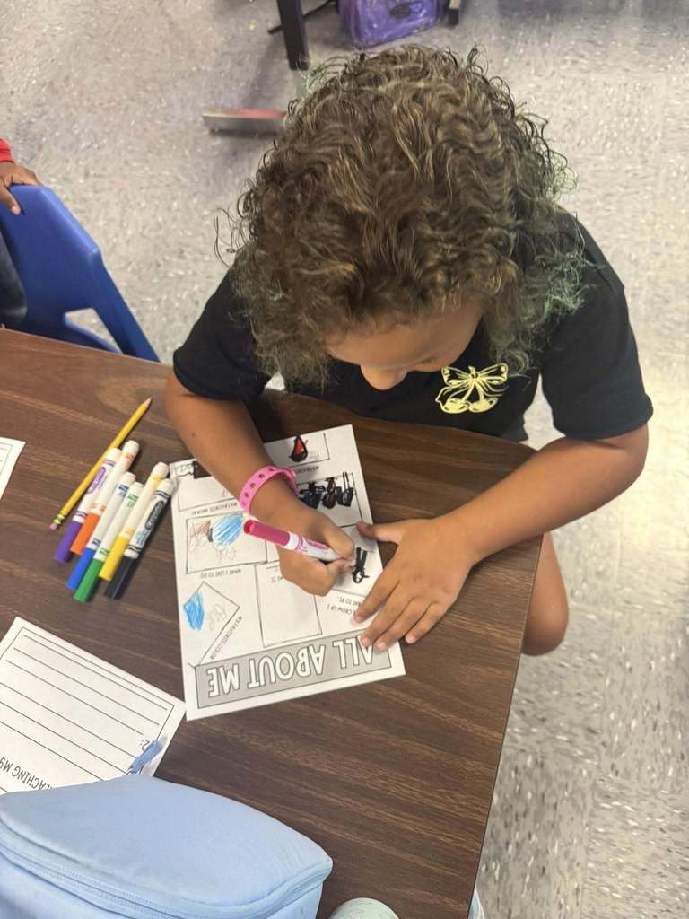 A young student with curly hair colors an “All About Me” worksheet at a classroom table with markers and pencils nearby.