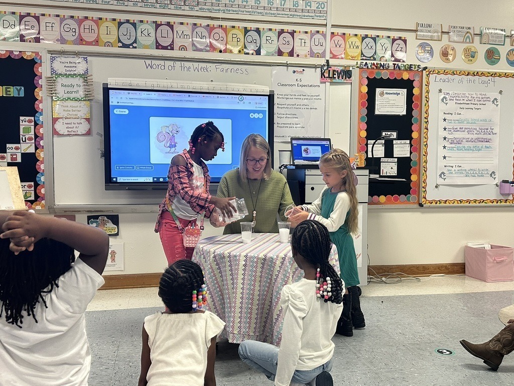 A teacher and two young students lead a classroom activity while classmates watch from the floor.