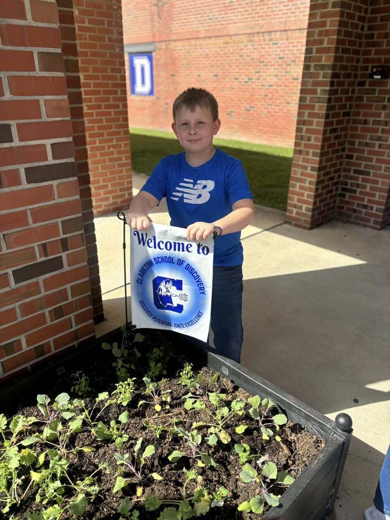 A student stands beside a raised garden bed outside, holding a “Welcome to Clarkton School of Discovery” sign.