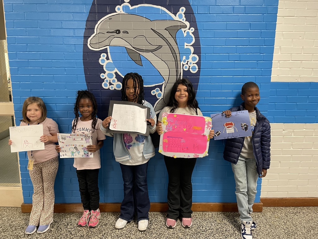 Five elementary students stand in front of the blue dolphin mural wall, each holding their creative drawings and posters. Their work includes colorful illustrations and words showing classroom learning and personal expression.