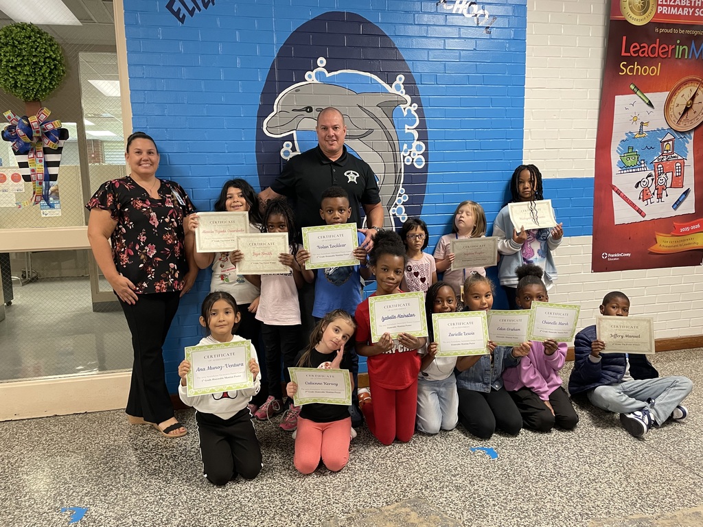A group of elementary students stand with two adults in front of a blue wall mural featuring a dolphin and the words “Elizabethtown Primary School.” The students proudly hold up certificates recognizing their achievements. The adults, one in a floral top and one in a black shirt with a badge logo, smile beside them.