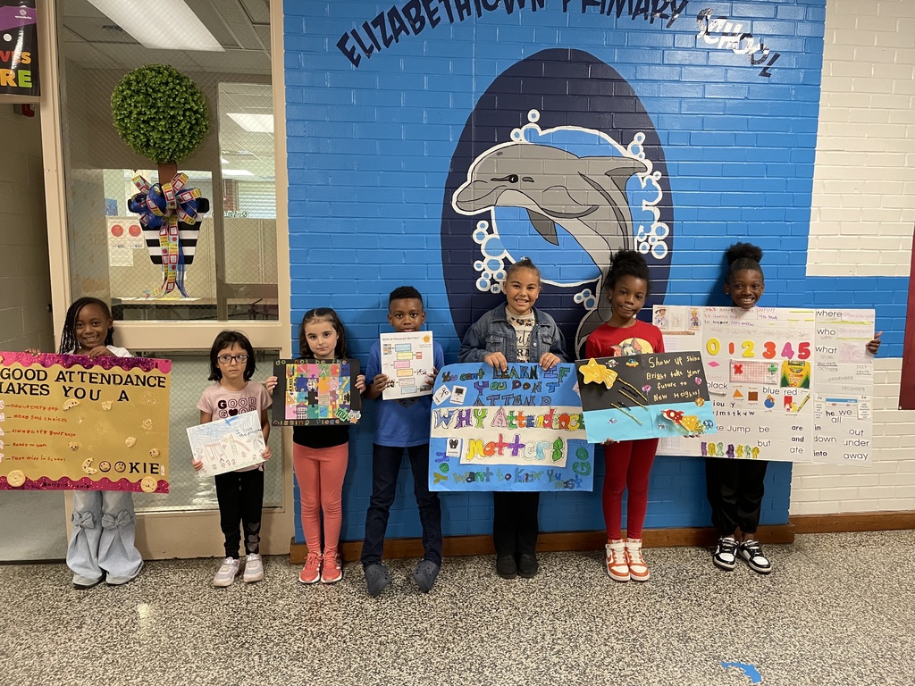Seven elementary students stand in front of a blue dolphin mural wall holding colorful posters they created about good attendance. Each poster is unique, featuring bright colors, drawings, and encouraging messages about why attendance matters.