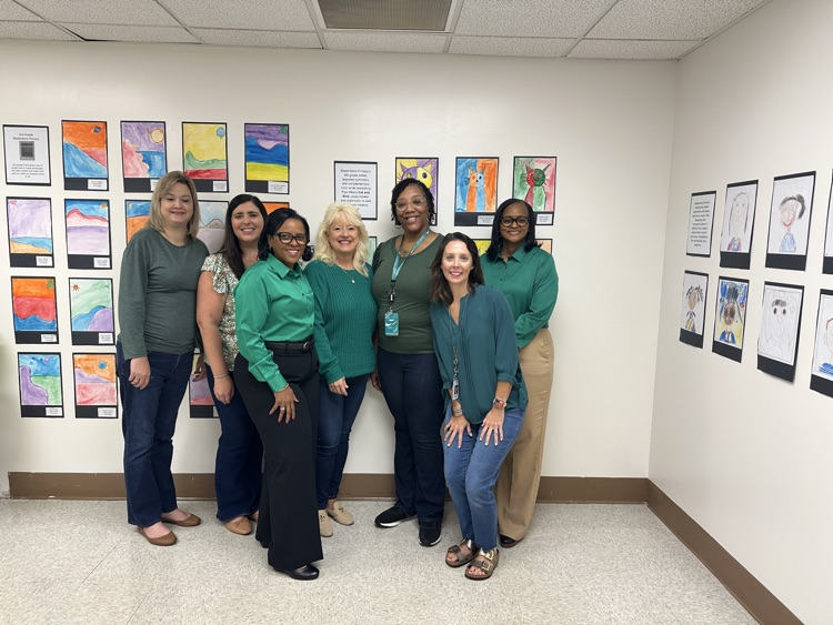 Seven Bladen County Schools district office staff members stand together in a hallway wearing various shades of green in recognition of World Mental Health Day. Behind them, colorful student artwork is displayed on the wall.