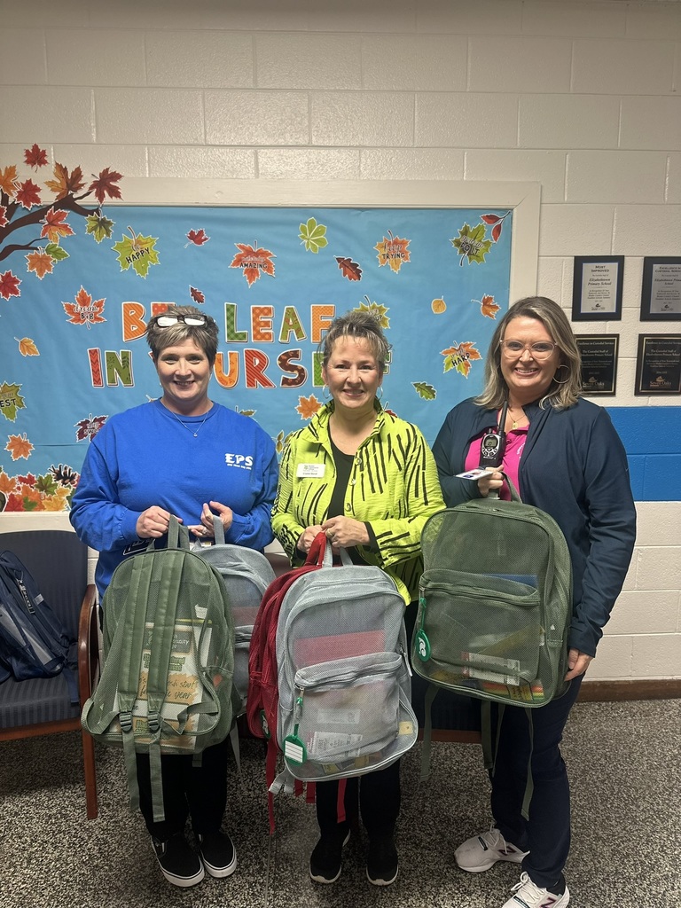 Three women stand smiling, each holding backpacks filled with supplies. A colorful "Believe in Yourself" bulletin board decorated with autumn leaves is behind them, adding a cheerful and motivational ambiance.