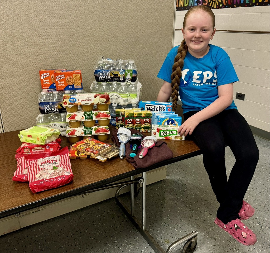A young girl in a blue shirt sits smiling next to a table filled with snacks, bottled water, and toiletries, conveying a sense of generosity and community.