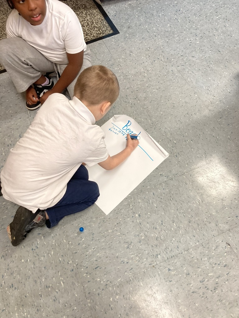 Two students sit on the floor working together on a large sheet of paper labeled “Beach.” One student writes ideas with a blue marker while the other looks on.