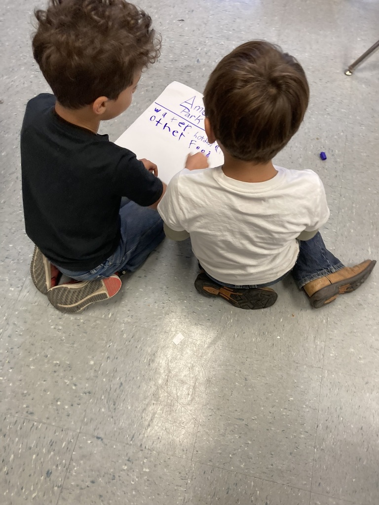 Two young students sit side by side on the floor writing on a paper labeled “Amusement Park,” brainstorming things like water bottles and food.