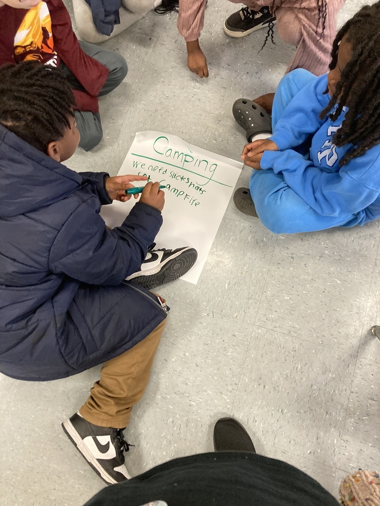 A small group of students sits in a circle on the floor. One student writes “Camping” on a poster and lists items they might need, like sticks and snacks.
