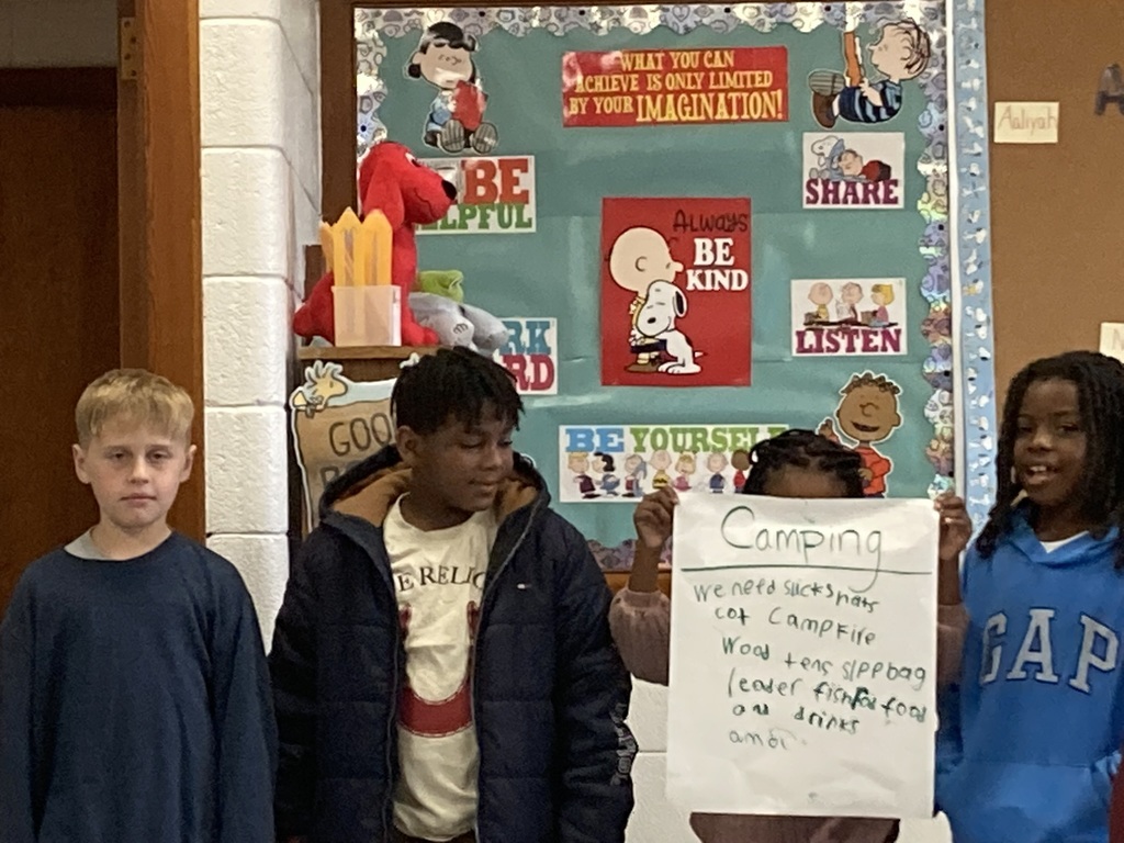 A group of four students stands together holding up their “Camping” poster and sharing their ideas in front of a colorful classroom bulletin board decorated with Snoopy and Charlie Brown sayings like “Be Kind” and “Be Yourself.”