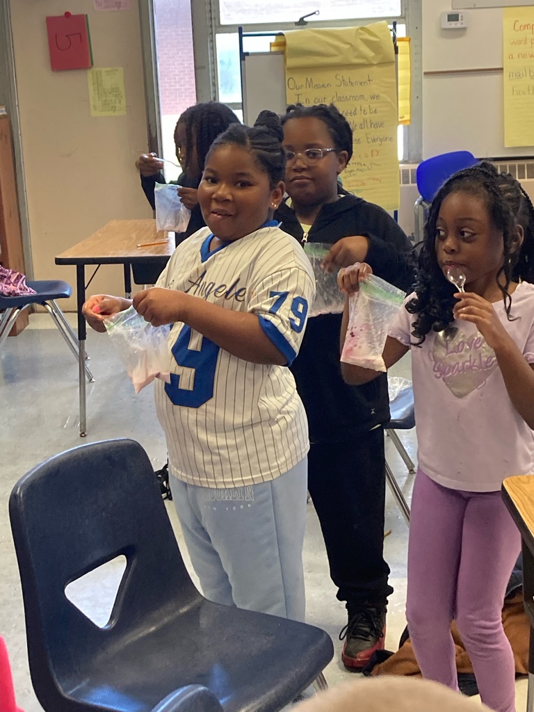 Three students stand proudly holding their completed ice cream bags and enjoy tasting their creations with spoons. The classroom background includes posters, charts, and a bright, cheerful atmosphere.