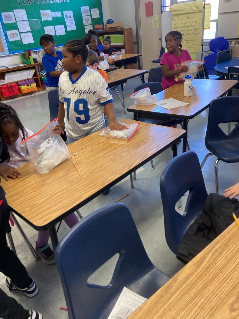 Several students stand and shake plastic bags filled with ingredients at their desks during the “Ice Cream in a Bag” experiment. They appear excited and engaged as they participate in the hands-on science activity.