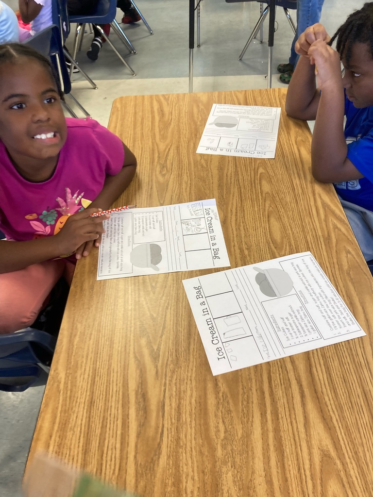 Two students sit at a classroom table smiling and looking at their “Ice Cream in a Bag” experiment worksheets, which include steps and pictures for the activity.
