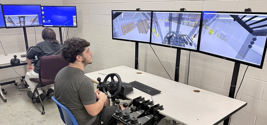 Two students sitting at simulation stations, each with a steering wheel, multiple control levers, and three large monitors displaying a 3D warehouse forklift simulator. The students are practicing operating virtual forklifts in a classroom with white brick walls.