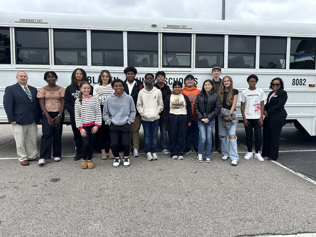 A group of sixteen high school students and two adults stand in front of a white Bladen County Schools bus in a parking lot. The students are smiling and dressed in casual clothing, while the adults on each end of the group are wearing professional attire. The sky is overcast, suggesting a cool or cloudy day.