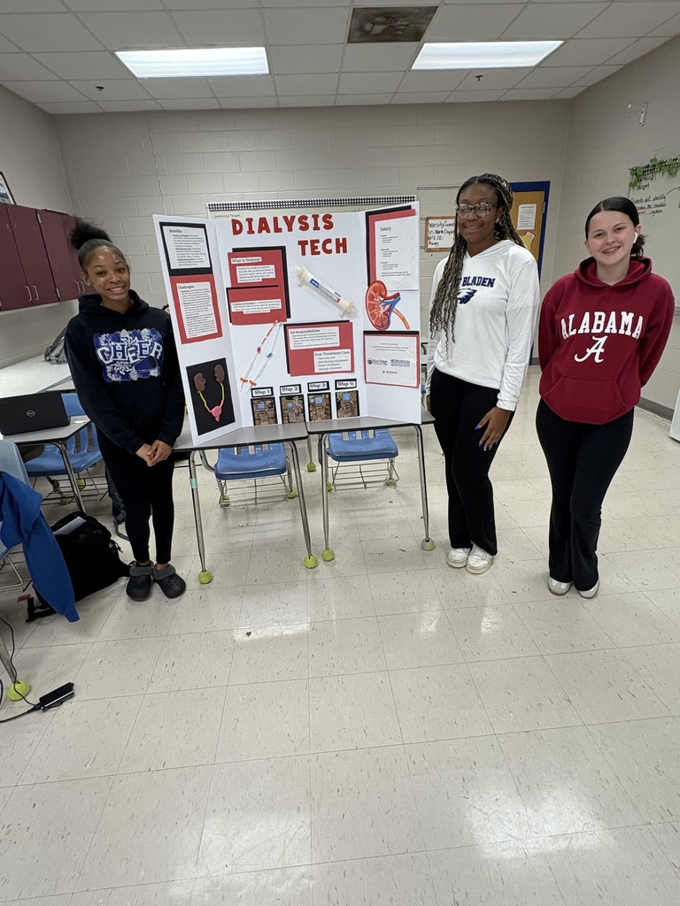Three students standing in a classroom beside a trifold presentation board titled ‘Dialysis Tech.’ The board includes text, images, and diagrams explaining dialysis technology. The students are smiling and posing for the photo.