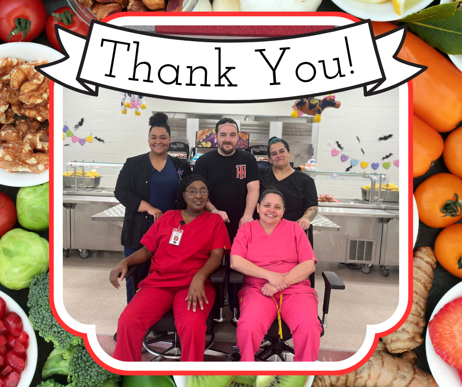 Five cafeteria staff members smiling together in the school cafeteria. Two are seated in front wearing red uniforms, and three are standing behind them in black attire. A banner above them reads “Thank You!” surrounded by colorful fruits and vegetables.