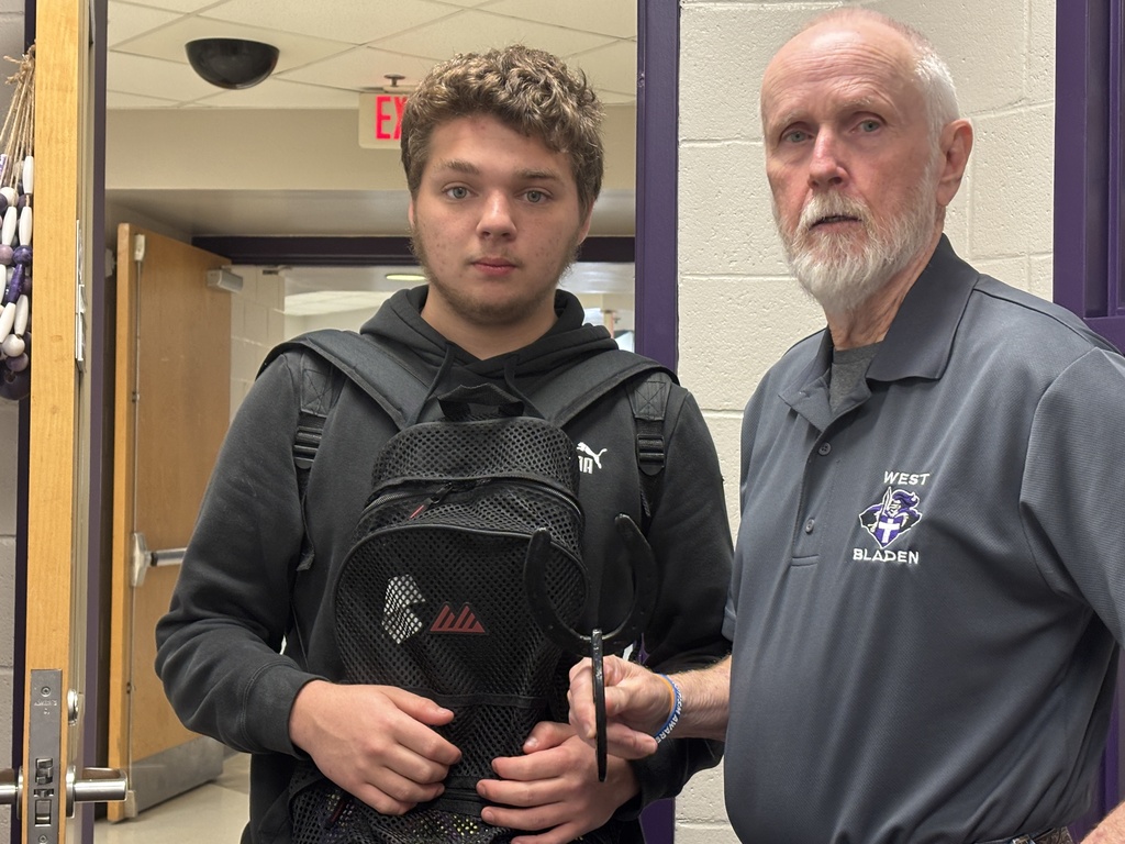 A teacher and a student at West Bladen High School stand next to a whiteboard with charts and maps. The teacher is holding a metal hat and jacket rack made from horseshoes, a project from the Agriculture Mechanics Honors class.