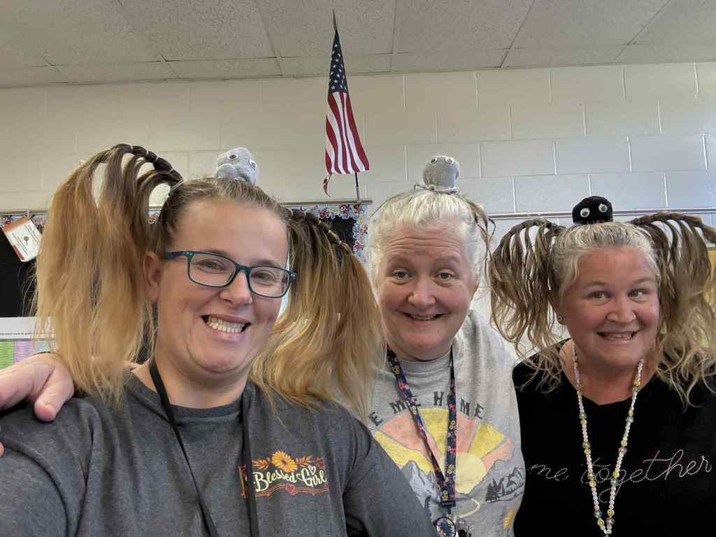 Three smiling teachers pose together in a classroom decorated for school activities. Each has creative, silly hairstyles with braided pigtails sticking out to the sides and small stuffed creatures sitting on top of their heads. They are dressed in casual shirts with cheerful designs and name lanyards, standing in front of an American flag and classroom wall. The photo captures their fun spirit and enthusiasm as they get ready to welcome families to the school’s Fall Festival event.