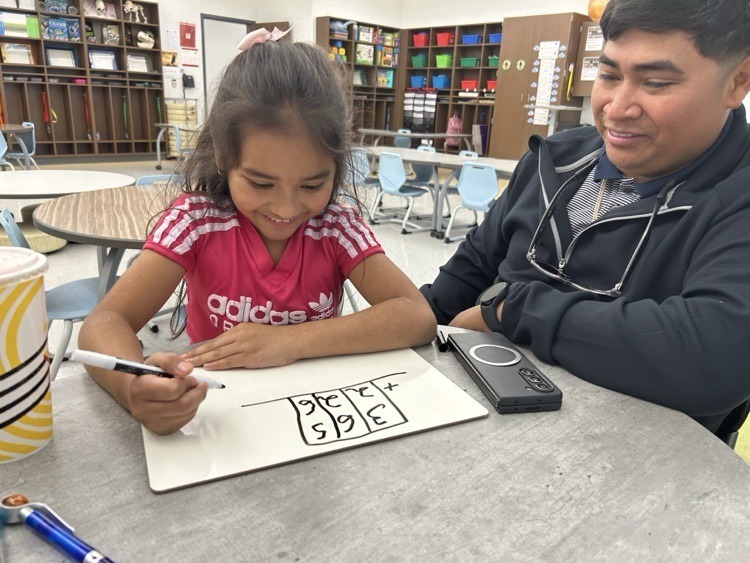 A young girl smiles as she works on a math problem written on a small whiteboard. An adult sits beside her, watching proudly.