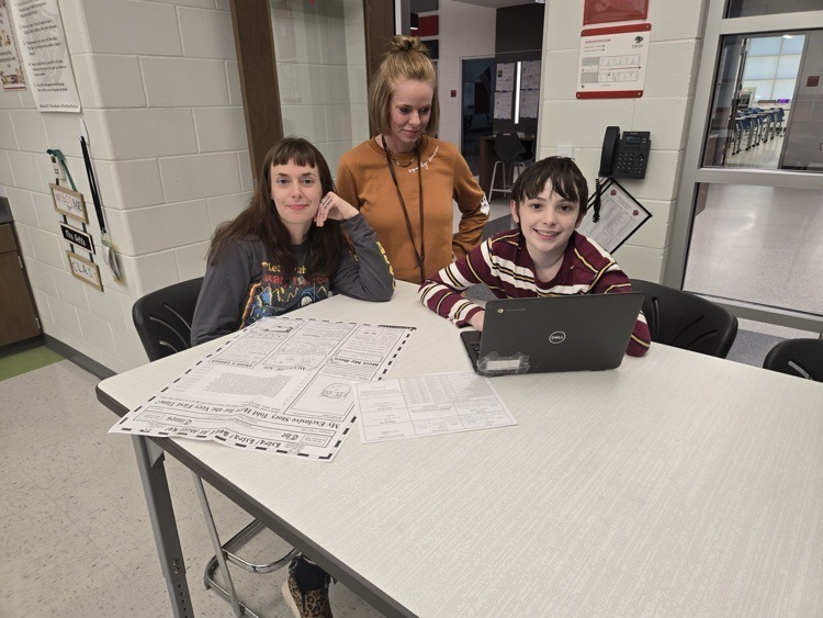 A student, teacher, and another adult sit together at a table in a classroom. The student uses a laptop while papers are spread across the table, and everyone is smiling.