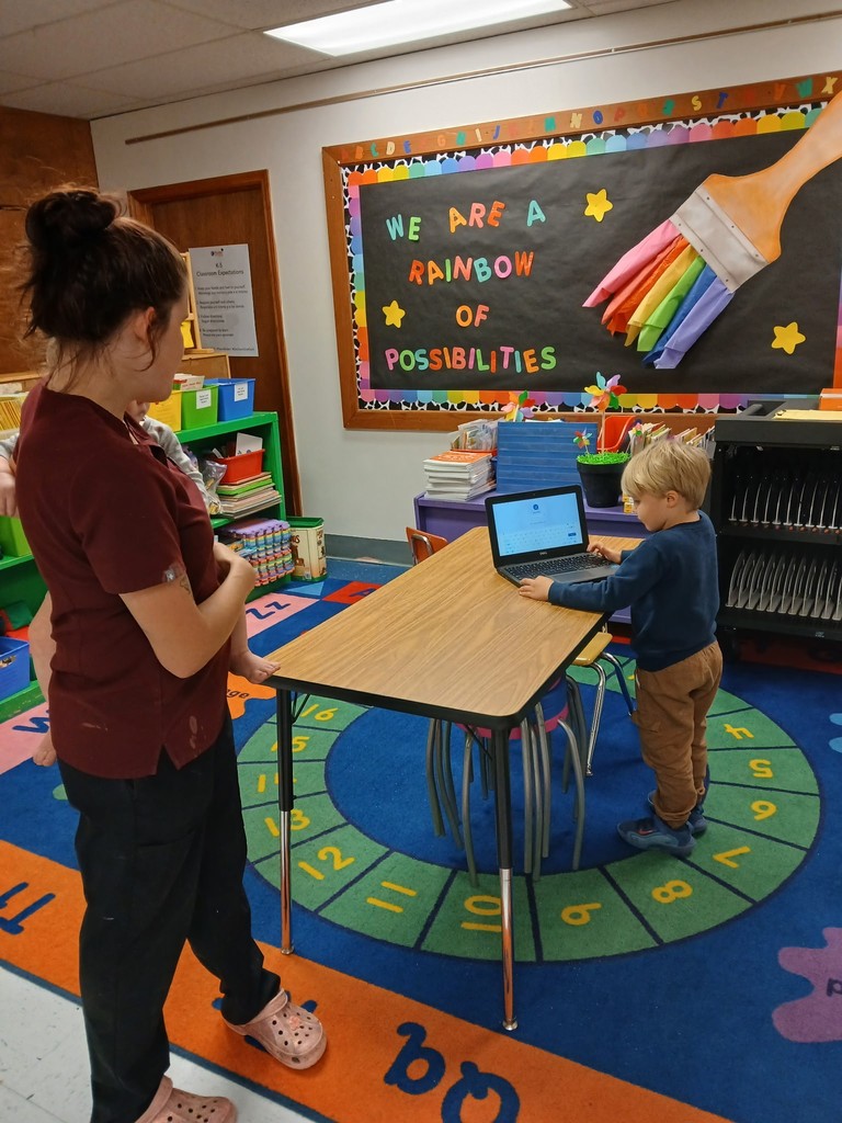 A young child stands at a small table using a laptop while an adult observes nearby in a colorful classroom with a “We Are a Rainbow of Possibilities” bulletin board.