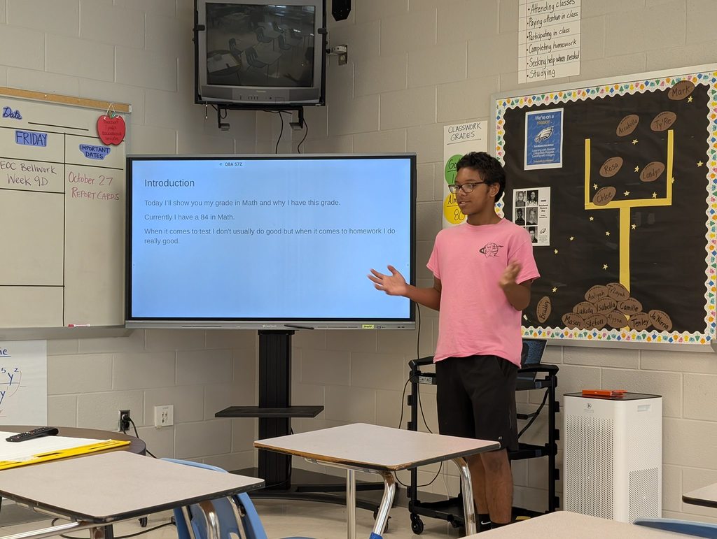 A student wearing a pink shirt presents in front of a classroom with a slide on the screen showing his math grade and reflection.