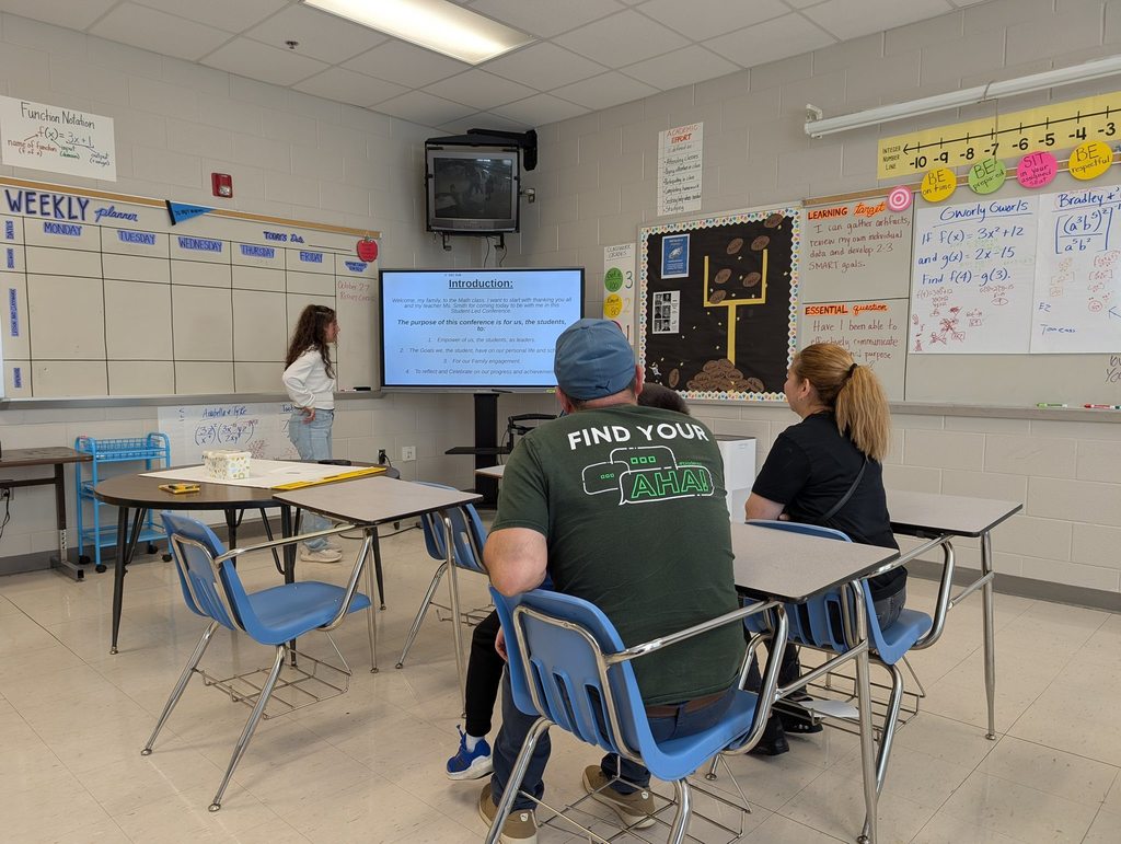 A student stands at the front of a classroom presenting to two adults seated at desks while a slide labeled “Introduction” is displayed on a large screen.