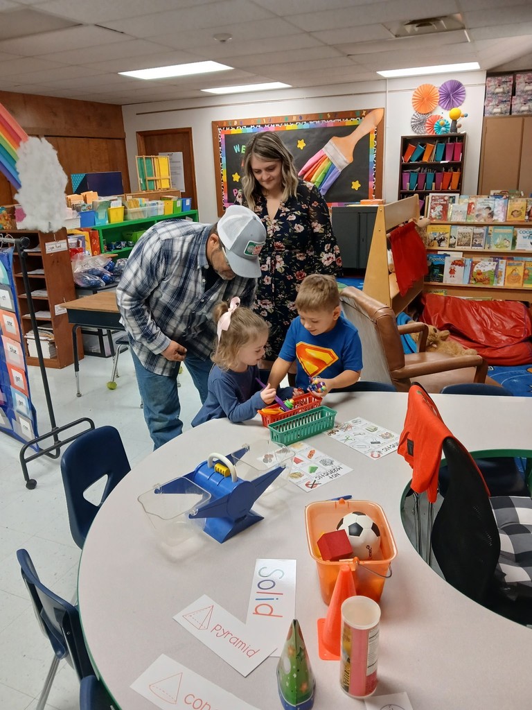 Two young children sit at a classroom table working on an activity while two adults watch and guide them in a colorful early learning classroom.