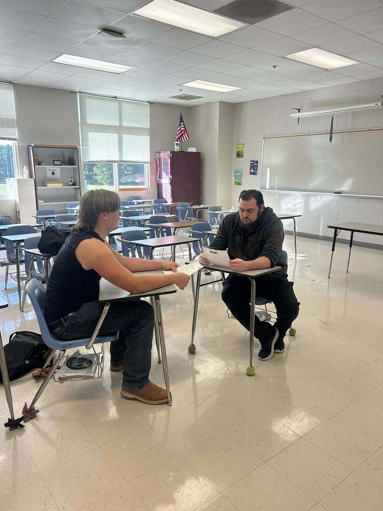 Two adults sit across from each other in a high school classroom discussing papers spread out on the desks.
