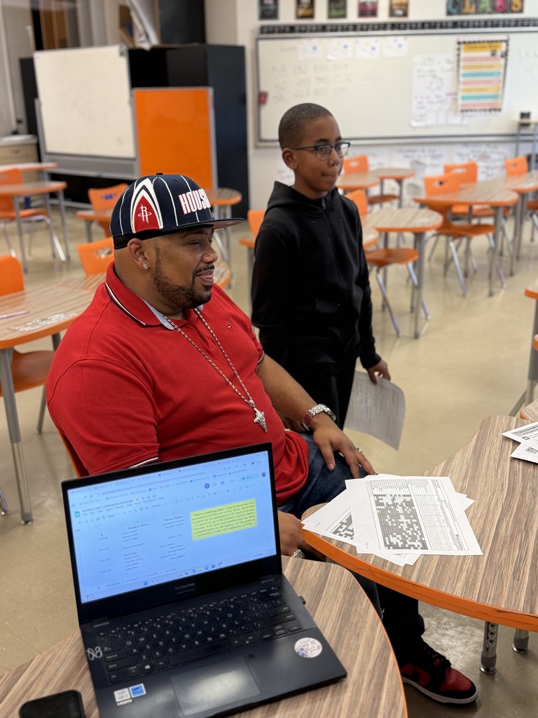 A student stands beside an adult seated at a desk in a classroom. The adult smiles as they review papers and a laptop open to a spreadsheet.