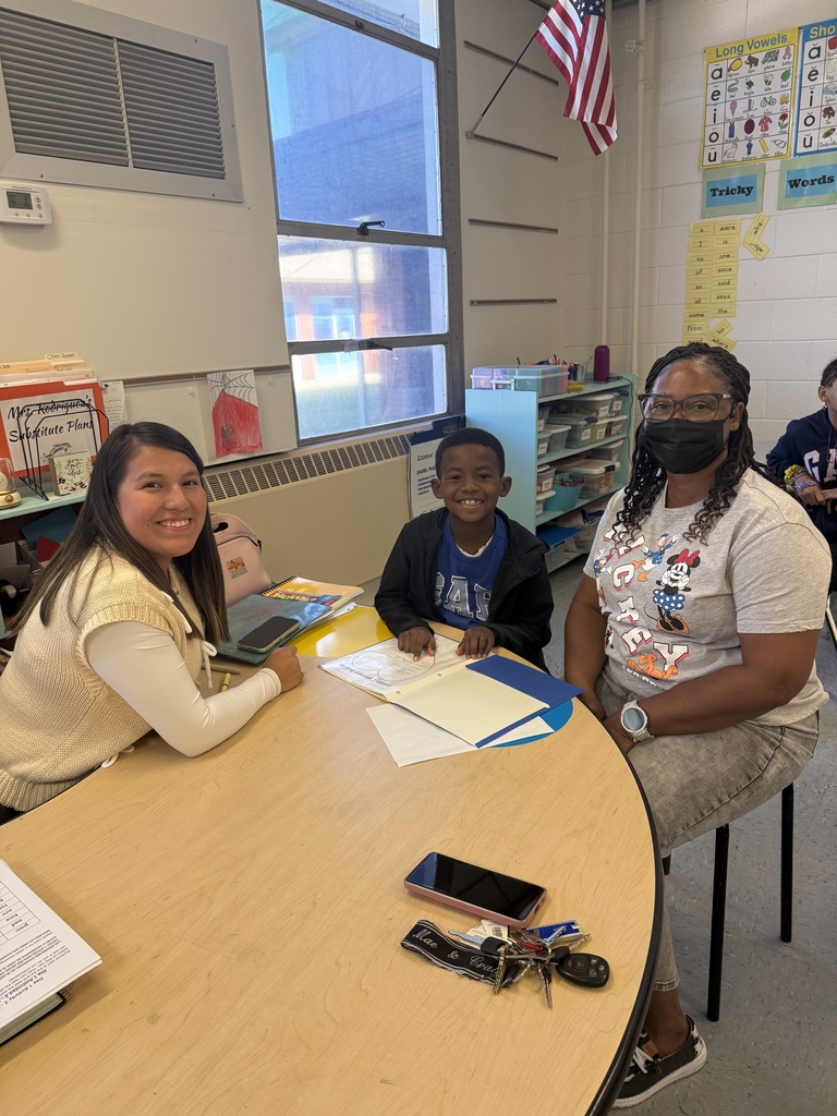 A young boy sits between two adults at a round classroom table smiling as they look over his folder and papers together.