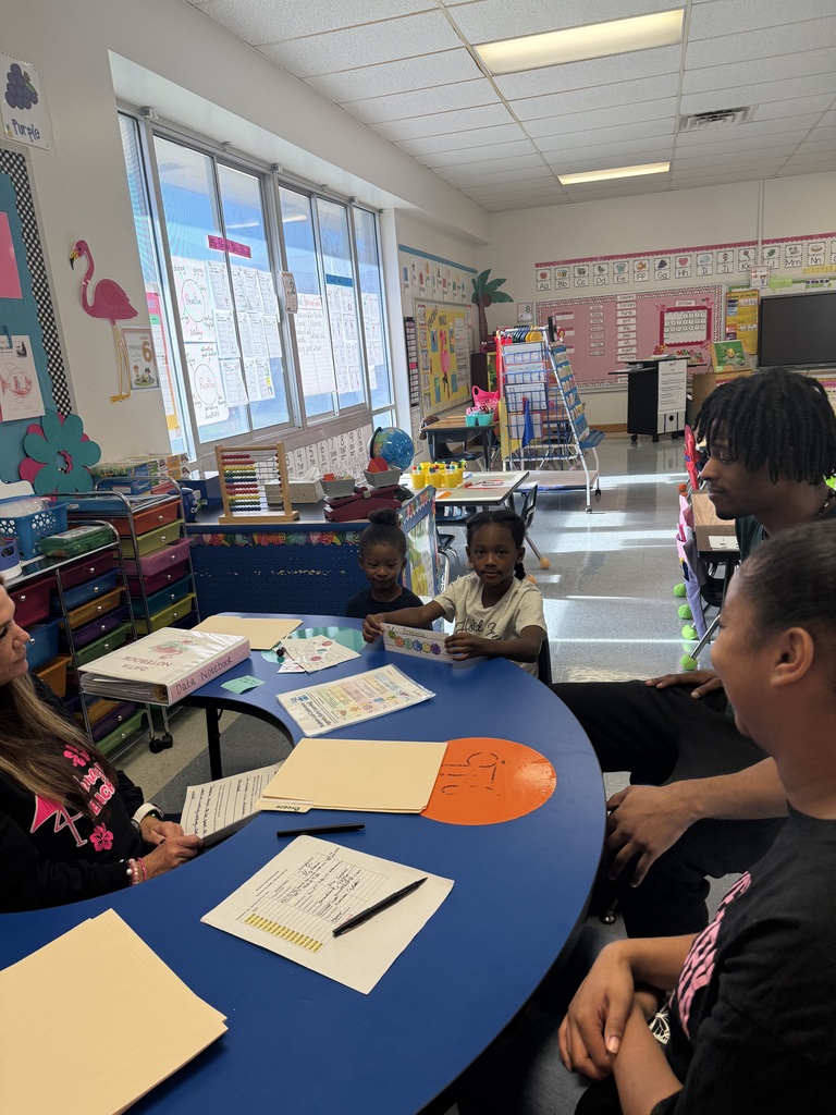 Two young students sit at a blue kidney-shaped table with two adults, sharing their progress sheets during a classroom conference.