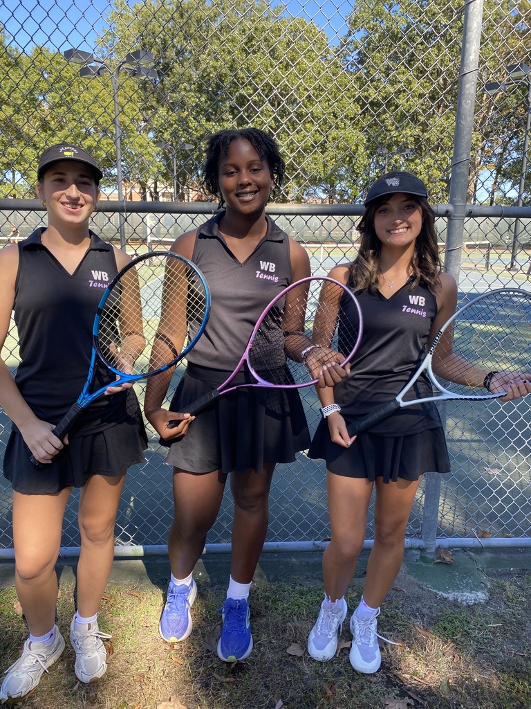 A group photo of four people standing in front of a tennis court fence. From left to right, a man wearing sunglasses, a hat, a gray jacket, and purple shirt stands beside three female tennis players wearing black WB Tennis uniforms and holding rackets. They are smiling and standing on grass with trees visible in the background.