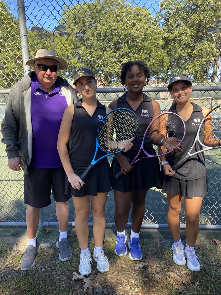 A group photo of four people standing in front of a tennis court fence. From left to right, a man wearing sunglasses, a hat, a gray jacket, and purple shirt stands beside three female tennis players wearing black WB Tennis uniforms and holding rackets. They are smiling and standing on grass with trees visible in the background.