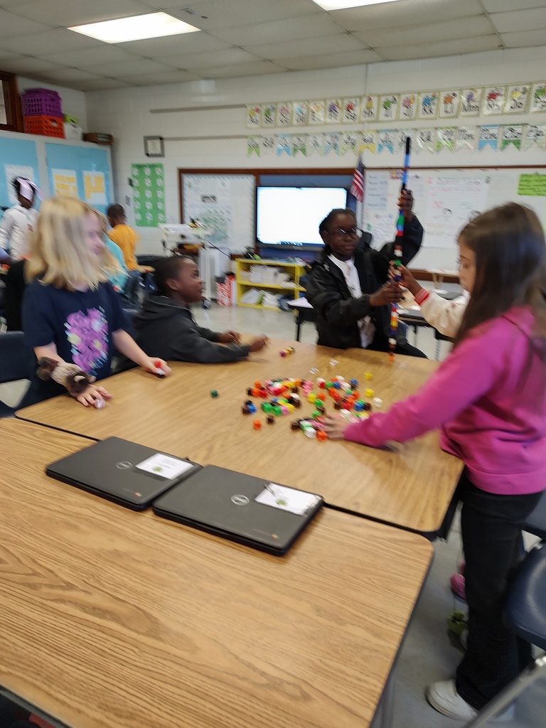 Each team collaborates, stacking cubes carefully and testing balance as they laugh and share ideas. The classroom is bright and cheerful, with charts, books, and student work displayed around the room.
