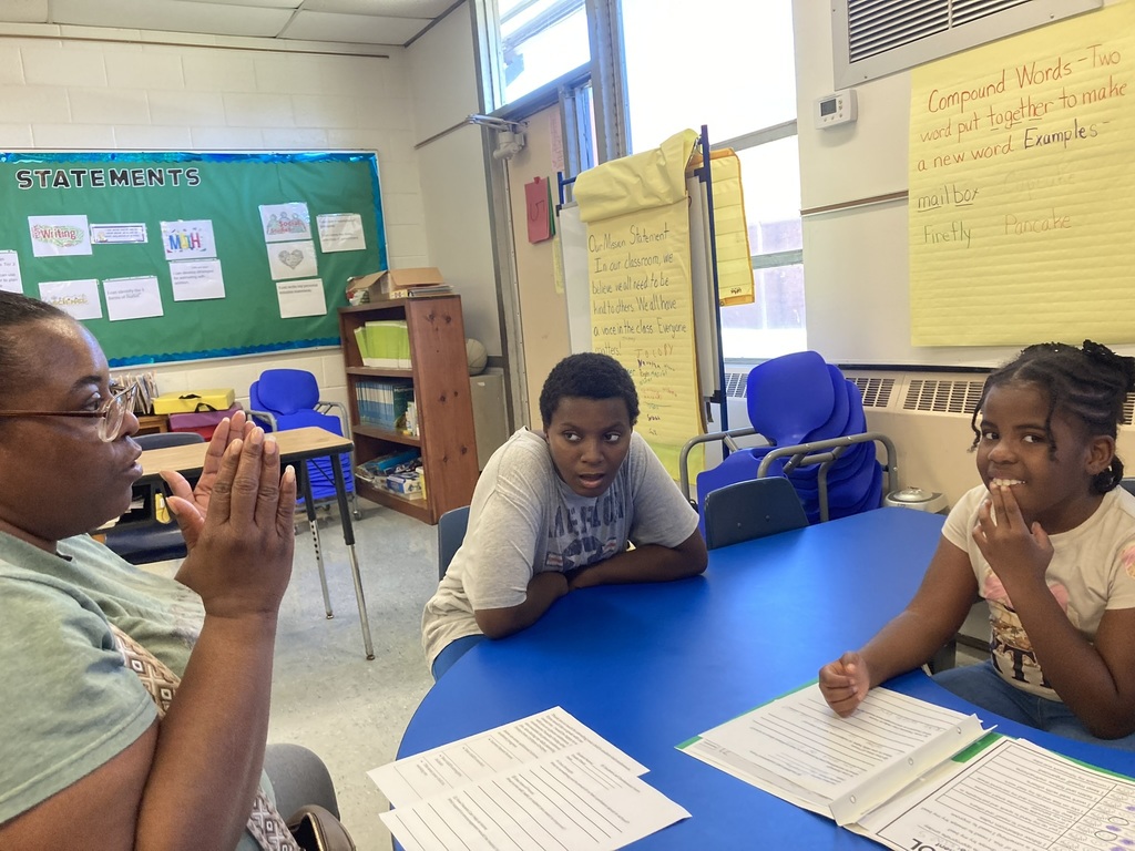 A teacher sits at a blue table with two students during a student-led conference. The teacher listens attentively with her hands clasped as one student smiles and another looks thoughtful. Papers with student work are spread out on the table, and classroom charts on the wall display a mission statement and examples of compound words.