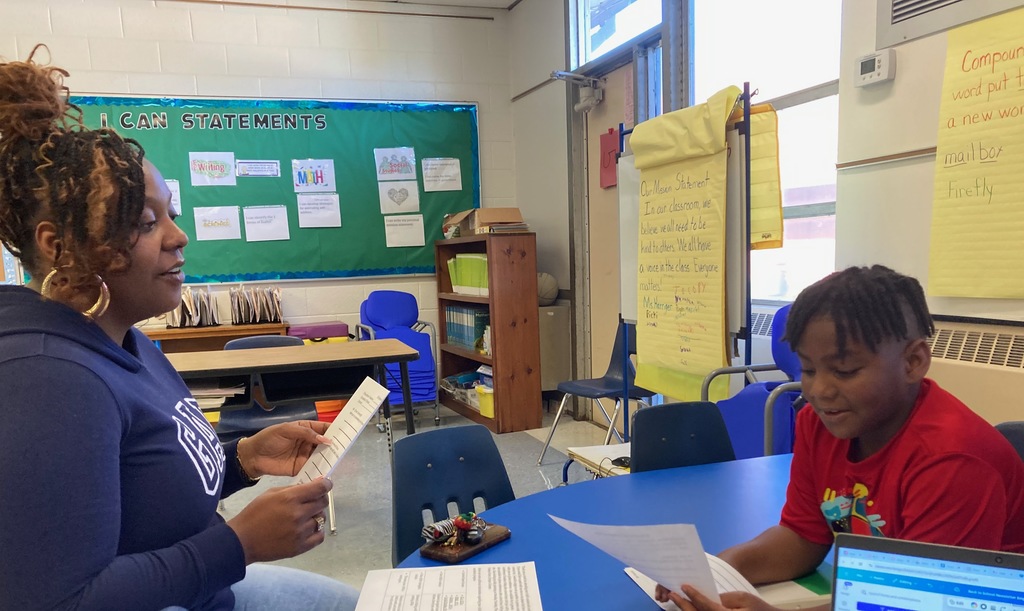 A student sits across from his teacher at a blue table, confidently reading from his paper during a student-led conference. The teacher smiles and listens while holding a copy of the student’s work. A laptop and classroom charts about writing and classroom goals are visible in the background.